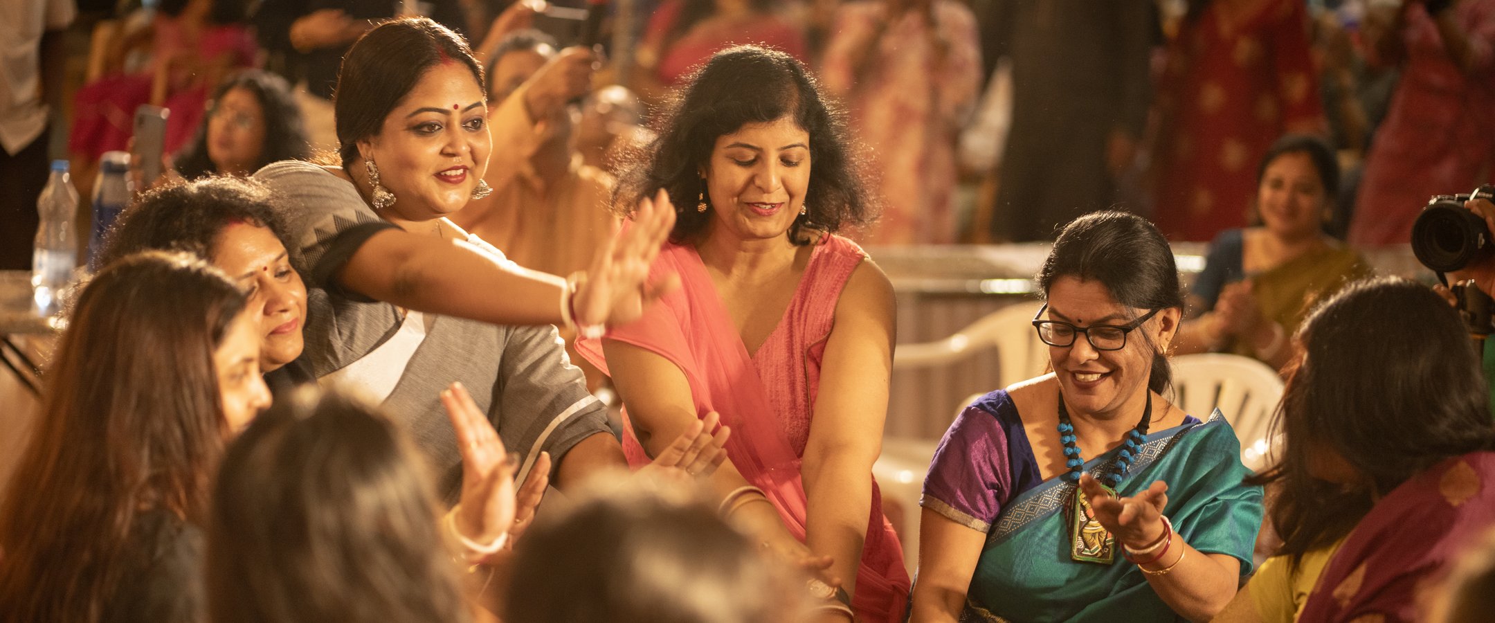 A circle of women in vibrant pink, teal, and grey sarees dance and laugh together at the Anandam Durga Pujo celebration — joy and movement fill the frame.