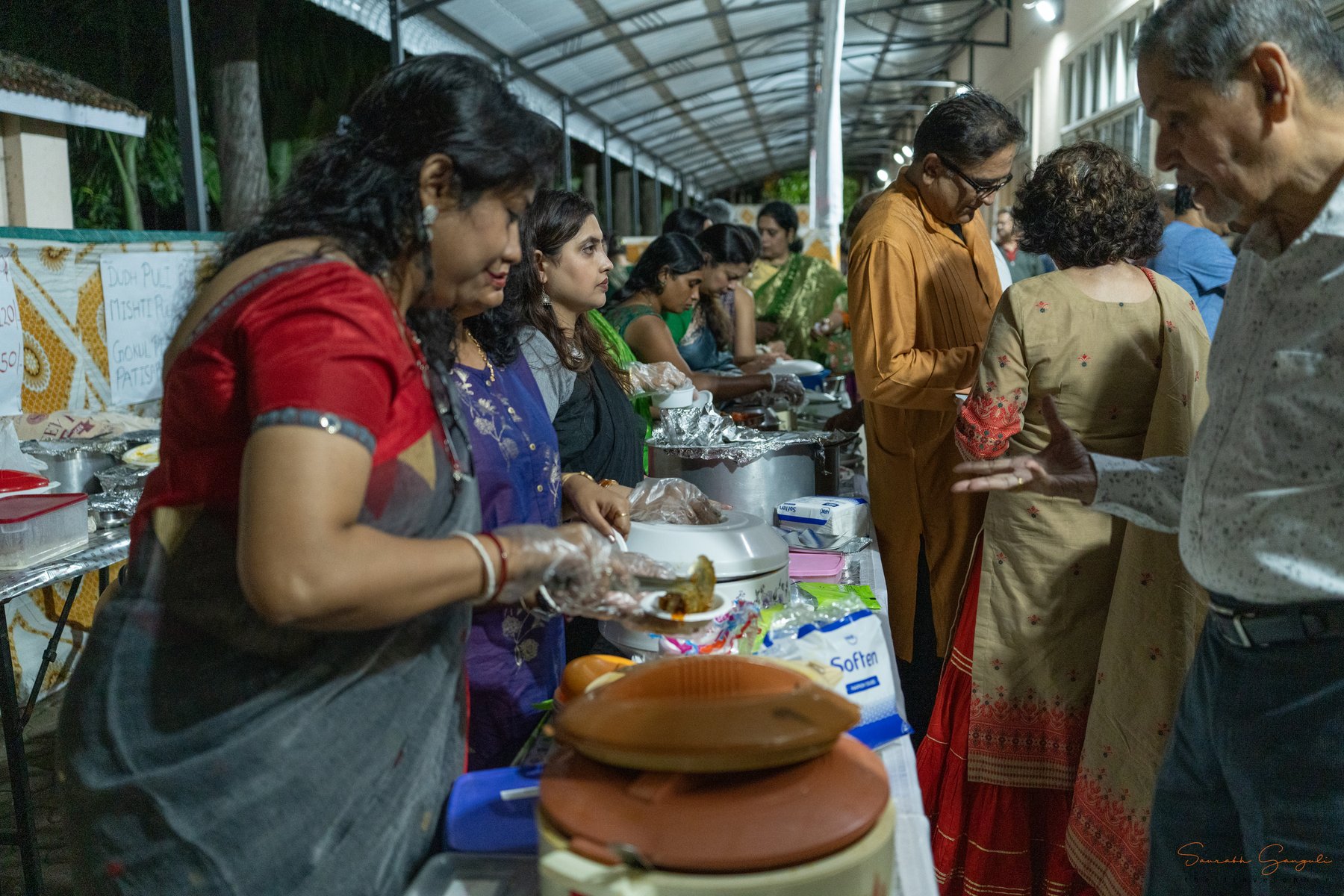 Volunteers in vibrant saris serve home-style Bengali fare from a stall at the Anandam puja — earthen pots, hand-written menu boards, and visitors gathered around.