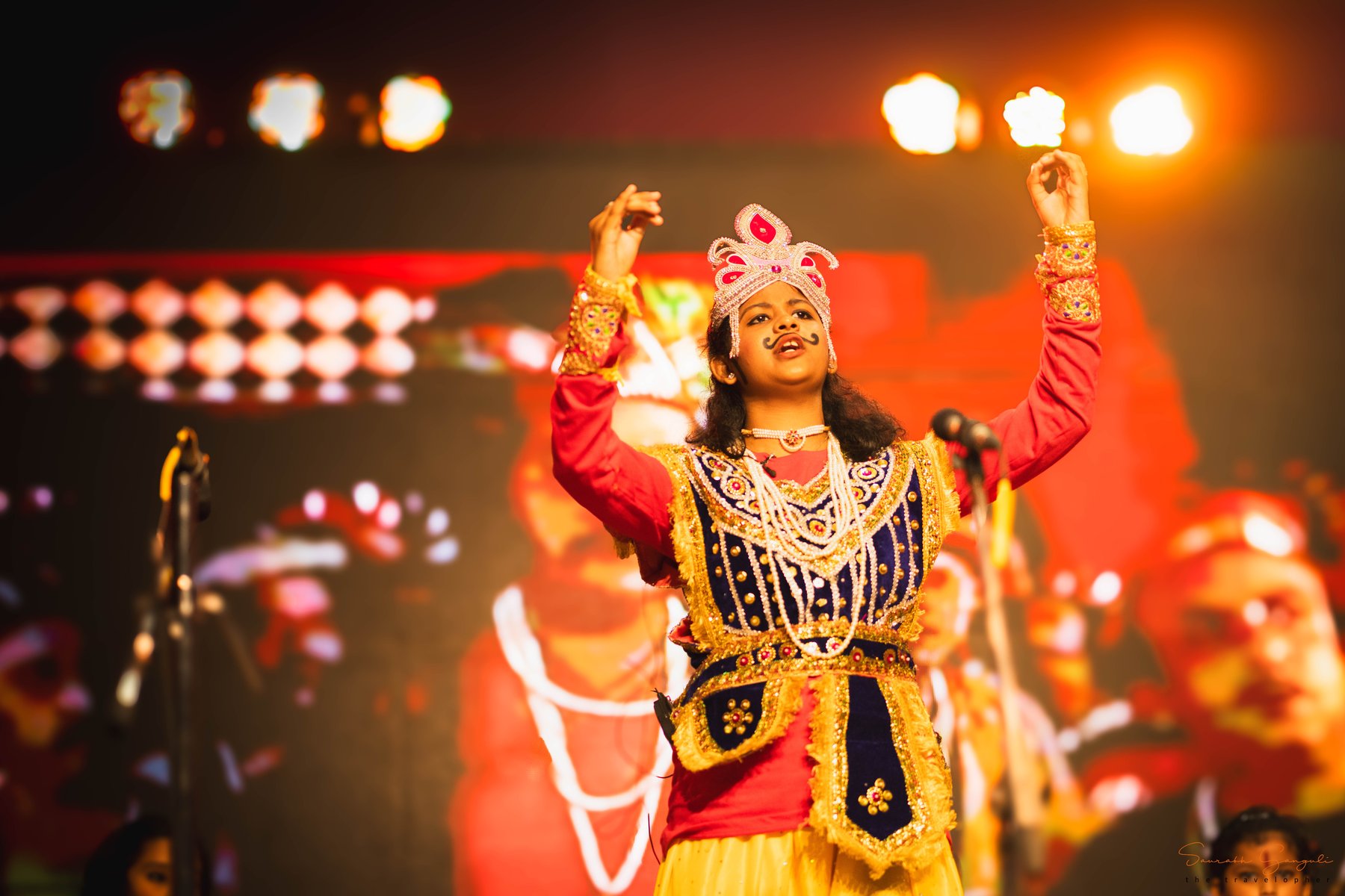 A young Anandam performer on stage in an ornate red-and-gold costume with a jewelled crown, arms raised mid-performance under warm stage lights.