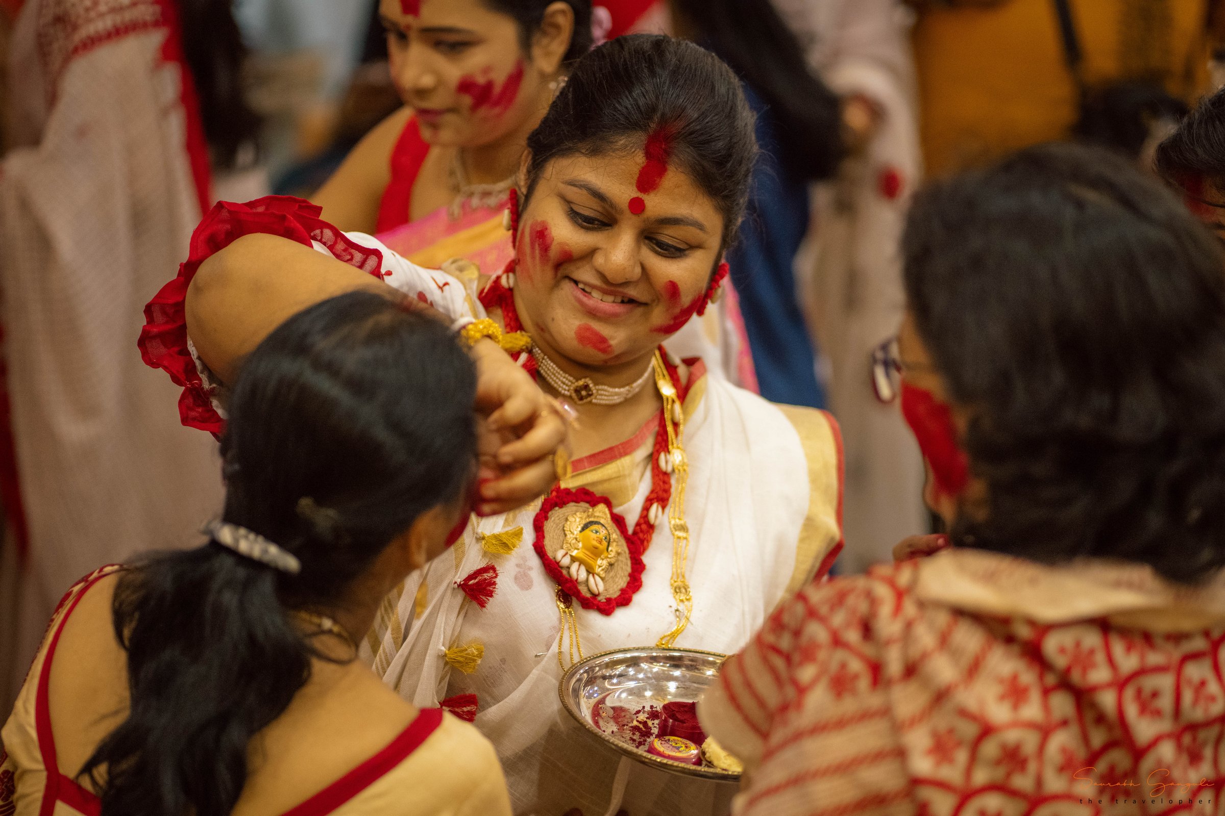 Women in white-and-red Bengali sarees applying sindoor to each other during Sindoor Khela — a Bijoya Dashami ritual.
