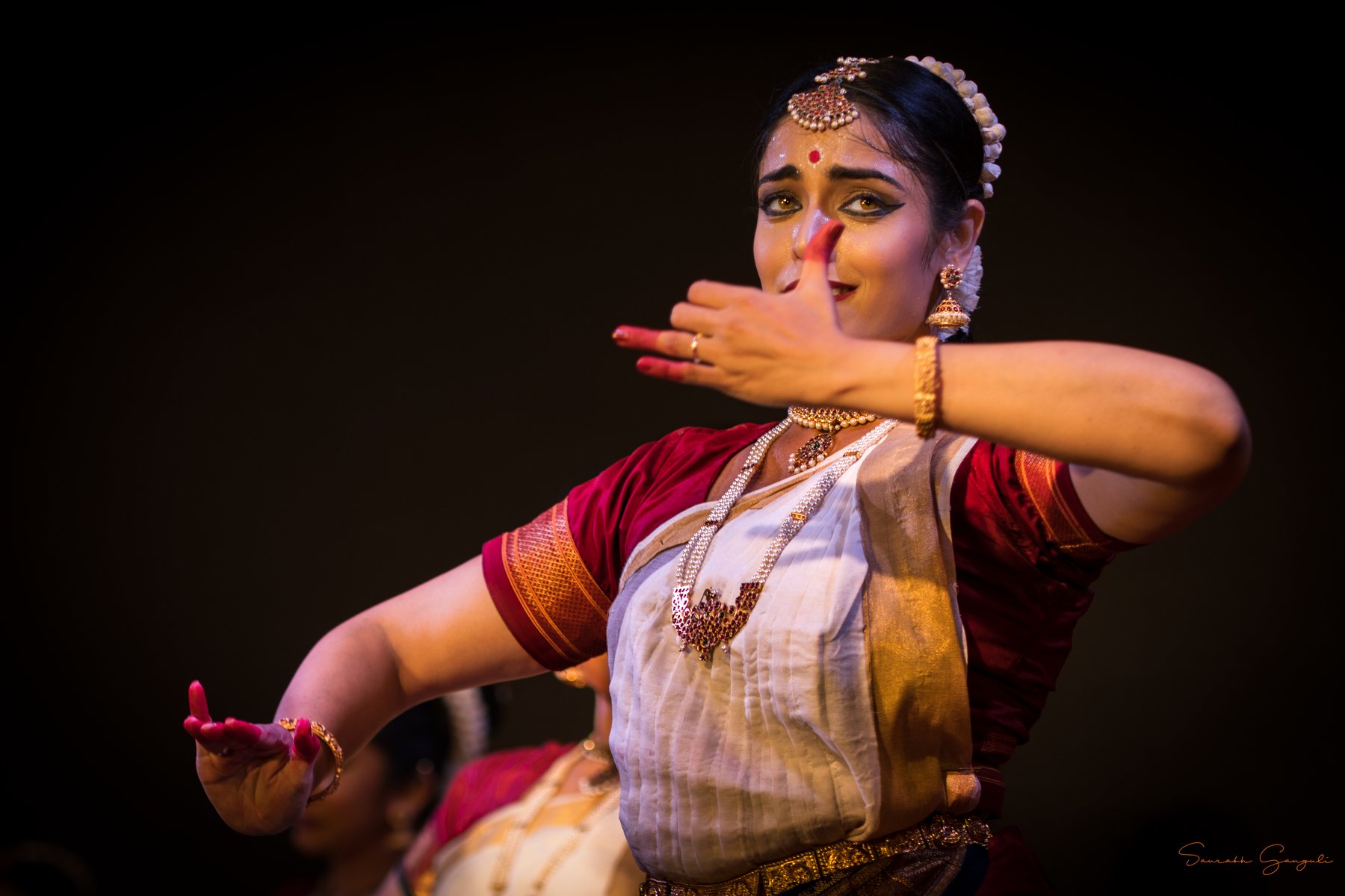 A classical Bharatanatyam dancer in a red-white-and-gold sari forms a graceful mudra, eyes expressive under stage lights.